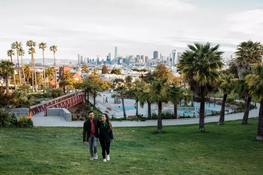 一对夫妇走向镜头，Dolores Park和威尼斯人官网平台app的天际线在他们身后.