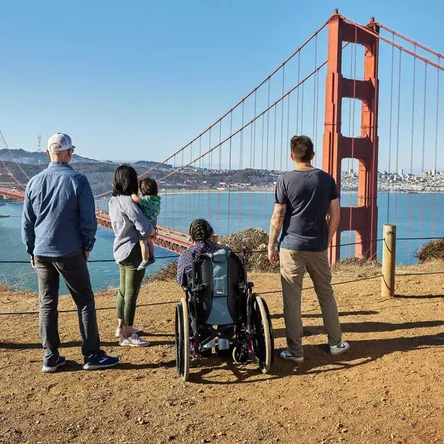 一群人, 包括一个坐轮椅的人, 从后面看, während sie von den Marin Headlands aus auf die Golden Gate Bridge blicken.